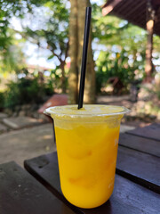 Orange ice is served in a plastic glass with a black straw, placed on a wooden table outdoors.
