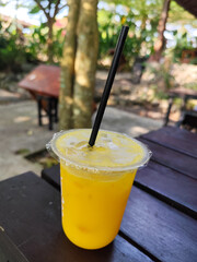 Orange ice is served in a plastic glass with a black straw, placed on a wooden table outdoors.