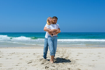 Couple standing barefoot on sandy beach with man hugging woman from behind in joyful moment.