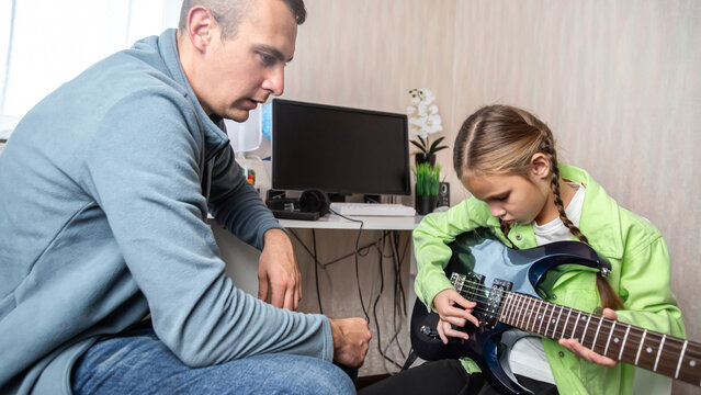 Girl practicing playing electric guitar and her father teaching her, spending a music lesson while sitting on the sofa at home.