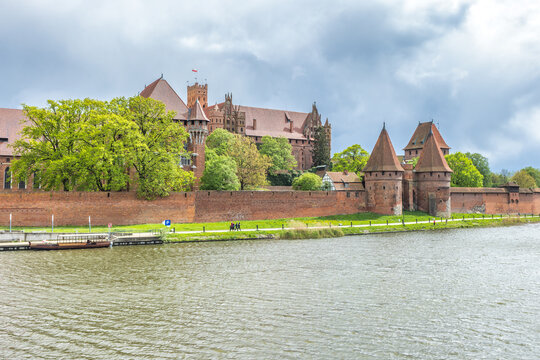 The Malbork Castle, a brick Gothic castle in northern Poland, Europe. Majestic medieval castle with red brick walls reflected in river under cloudy skies, showcasing historical architecture. - Powered by Adobe