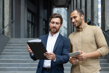 Two businessmen collaborating outdoors while reviewing documents and using a tablet device