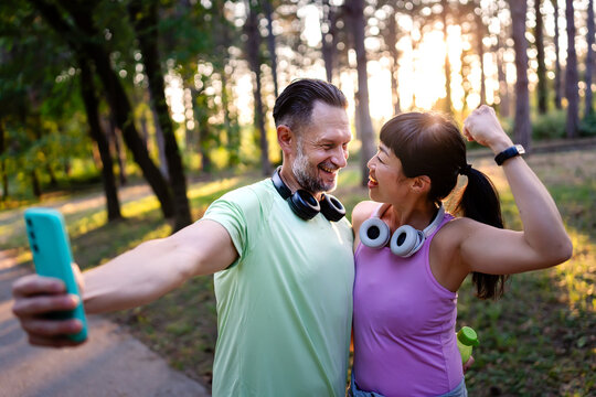 Happy Asian woman and Caucasian man taking selfie after outdoor workout in park at sunset, celebrating success and showing diverse friendship and active lifestyle