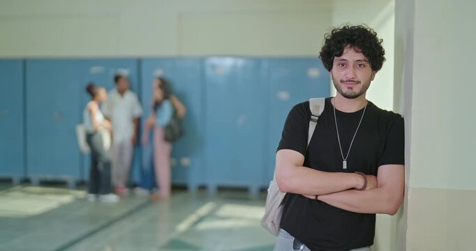Indian happy young adult gen z proud man male look camera cross arm standing campus area. Cool pupil boy curly hair wear bag fold hand staring cam do casual pose enjoy new good day time indoor place