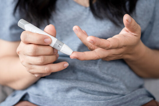 Asian woman at home checking her blood sugar by using a digital lancet on her finger and monitoring glucose levels with a glucose meter, showing diabetes care, self-testing