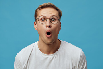 Surprised man with glasses wearing casual white T-shirt looking shocked in front of bright blue...