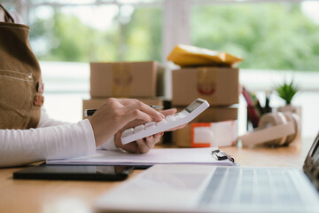 A hardworking young woman in an apron handles online orders, multitasking from packing boxes to...