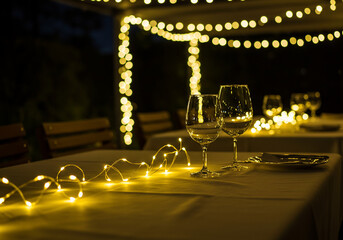 Obraz premium Two wine glasses stand on a table decorated with string lights under a canopy illuminated by warm, glowing fairy lights at night.