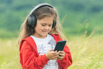 Cute little girl in wireless headphones holding smartphone and enjoying music outdoors