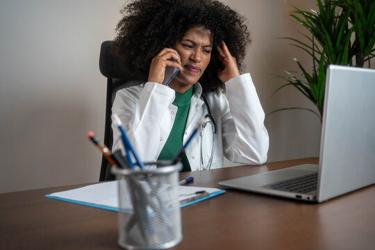 Stressed young female doctor talking on phone while working on laptop in office, multitasking under pressure - Powered by Adobe
