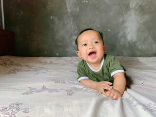 Fototapeta premium A happy baby boy is lying on his stomach, propped up on his hands, and looking directly at the camera with a wide, joyful smile. He is wearing a simple green t-shirt with white trim.