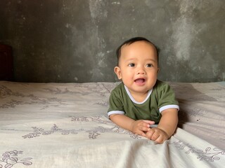 Fototapeta premium Cute baby boy lying on his stomach on the bed, smiling happily in natural light with a plain wall background.