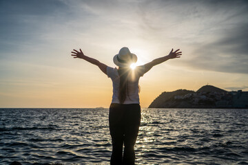 Mujer viajera con sombrero con los brazos abiertos hacia el sol en el horizonte marino,...