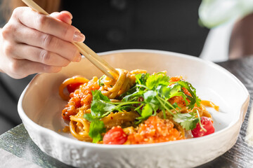 Close-up of hand holding chopsticks with noodles in bowl, garnished with cilantro and tomatoes....