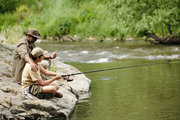 Middle aged man sitting beside boy fishing together on rocky riverbank, man giving encouraging...
