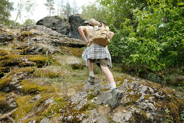 Boy hiking up rocky hillside with backpack, climbing steep terrain surrounded by green trees and...