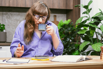 Elderly business woman drinking coffee from blue cup while working at desk in stylish modern...