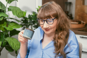 Elderly woman drinking coffee from blue cup while working at desk in stylish modern apartment