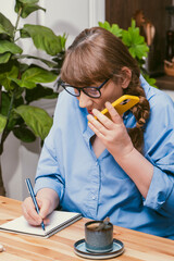 Serious elderly woman talking on smartphone and makes notes in notebook in stylish modern flat