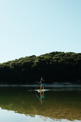 Naklejka premium Man paddleboarding on calm lake water surrounded by forest, enjoying peaceful outdoor activity in nature on a sunny day