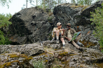 Middle aged Black man and teenage boy sitting on rocky terrain holding fishing rods and map, engaging in outdoor activity together surrounded by natural landscape
