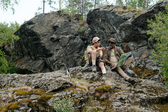 Middle aged Black man and teenage boy sitting on rocky terrain holding fishing rods and map, engaging in outdoor activity together surrounded by natural landscape