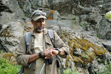 Middle aged man standing outdoors near rocky terrain checking wristwatch while holding fishing rod,...
