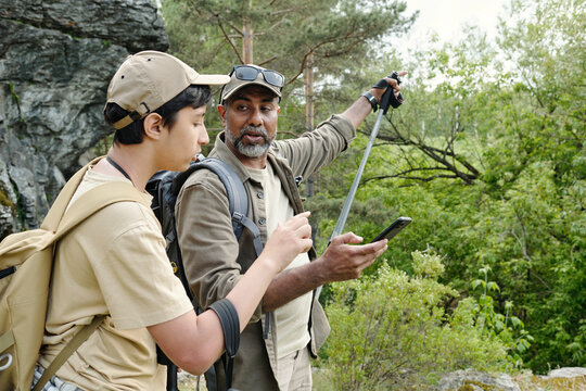 Middle aged South Asian man showing smartphone to teenage South Asian boy while standing outdoors with backpacks, both holding fishing rods, surrounded by green forest and rocky terrain