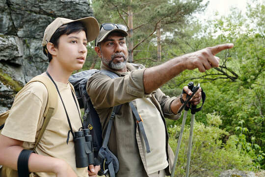Middle aged man and teenage boy standing outdoors in forest, man pointing into distance while boy watching attentively, both carrying fishing and hiking gear - Powered by Adobe