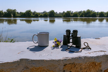 White hiking mug with bouquet of wild flowers and binoculars on half-ruined wall near lake, travel...