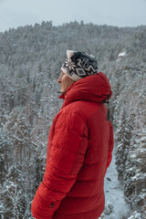 man stands on top mountain, against background an winter forest, wearing bright red puffer jacket...