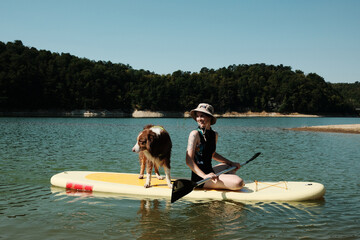 Woman and her Australian shepherd dog sitting on a paddleboard, smiling and enjoying a sunny day on...