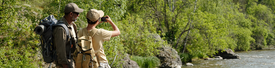 Middle aged man and boy standing on riverbank fishing together, both wearing backpacks, boy holding...