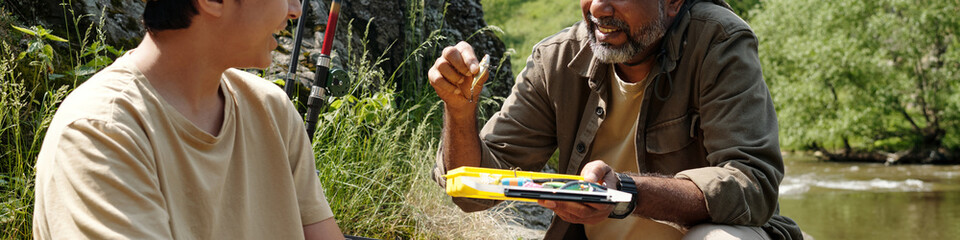 Middle aged Black man teaching fishing technique to Asian boy near riverbank, both sitting on grass...