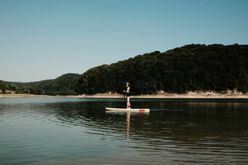 Woman paddleboarding on a calm lake with hills and trees in the background. Summer outdoor activities on water