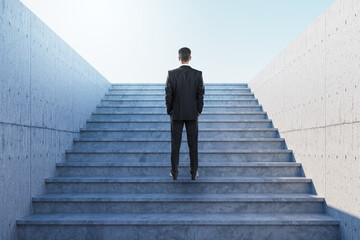 Businessman standing on modern staircase looking up toward bright sky, symbolizing ambition,...
