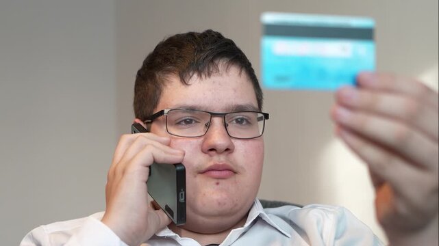 A teenage boy talks on a smartphone and reads a CVV number from a plastic card. deception telephone fraud concept