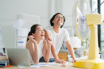 Air condition broke Hot asian female and male man in home studio sitting in front of fan Tired...
