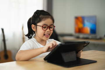 Cheerful Asian girl with glasses engaging with tablet computer at home in cozy environment,...