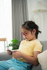 Young girl enjoying screen time, sitting comfortably on a sofa at home, wearing glasses and yellow...