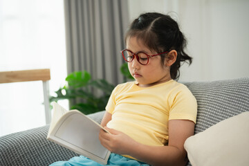 Young Girl with Glasses Reading a Book on a Cozy Sofa in a Bright and Modern Living Room...