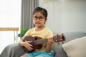 Young Girl with Glasses Playing Ukulele at Home in Cozy Living Room with Natural Light and Soft...