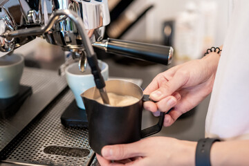 Barista frothing milk with steam wand in metal pitcher, espresso machine and cups visible. Coffee preparation in café or home kitchen