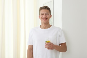 Smiling man with glass of water with cucumber and lemon near white wall indoors. Refreshing drink