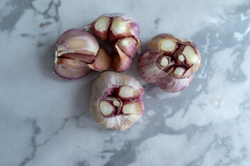 A close-up shot of partially peeled garlic bulbs on a light marble background. The visible cloves and purple-striped skin highlight the raw texture of this key ingredient, perfect for a food blog 