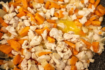 A close-up shot of chopped raw chicken and diced carrots being cooked with onions in a frying pan....