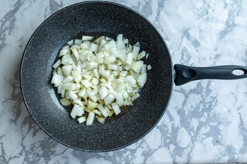  top-down shot of chopped white onions being sauteed in a dark, non-stick pan. The onions are...
