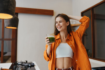 Young woman enjoying a green smoothie in a modern kitchen, wearing a bright orange shirt, exuding...