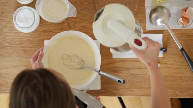A woman uses a culinary trick for thin crepes, pouring boiling water from a kettle into the pancake batter. This technique of scalding the dough helps to create a delicate, lacy texture. Top view.
