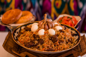 Traditional Uzbek dish pilaf, featuring fragrant rice, tender meat, and garnished with eggs, served on a decorative platter with bread and fresh vegetables. Copy space, menu